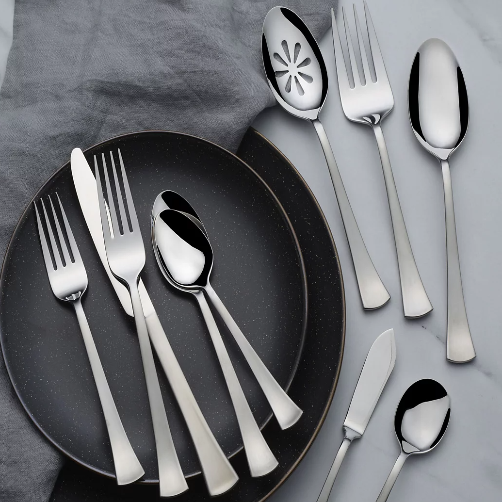 A set of stainless steel flatware includes forks, knives, and spoons arranged on two dark plates and a gray cloth.