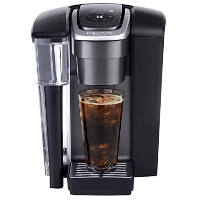 A single-serve coffee maker with a visible water reservoir, featuring an iced coffee brewing option and a tall glass filled with iced coffee on the drip tray.