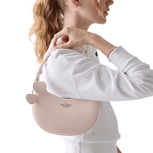 A person holds a light pink Kate Spade handbag with heart-shaped details on the strap, worn over a white sweatshirt.