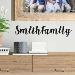 Wall decor with the text \“Smith Family\“ in black script sits above a wooden cabinet holding a plant, books, and decorative items, with a framed family photo above.