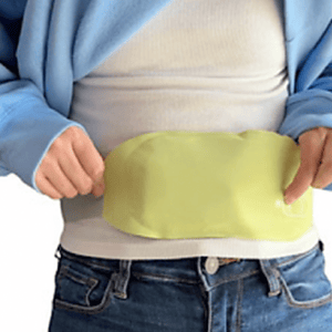 A person places a yellow-green portable heating pad on their lower abdomen. The pad is designed for pain relief and warmth, and its size and flexibility make it suitable for covering a range of body parts.
