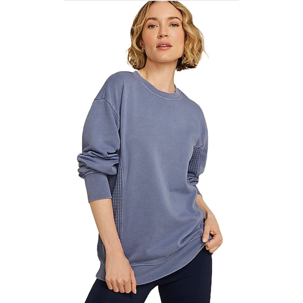 A woman wearing a loose-fitting, long-sleeved blue sweatshirt with ribbed cuffs and a waffle pattern detail on the sides, paired with dark leggings.