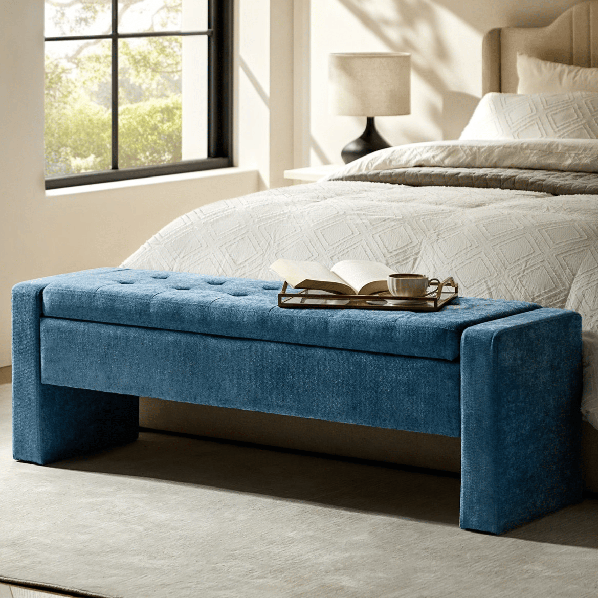 Blue upholstered storage bench at the foot of a bed with a tray holding a book and a cup, situated in a well-lit bedroom.