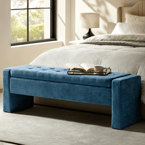 Blue upholstered storage bench at the foot of a bed with a tray holding a book and a cup, situated in a well-lit bedroom.