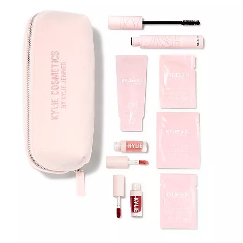 A pink cosmetic bag with a zipper contains mascara, skincare packets, a small tube of skincare cream, and two liquid lipsticks in light and dark shades. Packaging displays logos but avoid brand identification.