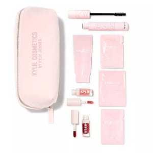A pink cosmetic bag with a zipper contains mascara, skincare packets, a small tube of skincare cream, and two liquid lipsticks in light and dark shades. Packaging displays logos but avoid brand identification.
