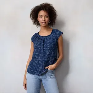 A sleeveless navy top with a pleated round neckline and an allover white polka dot pattern, worn with light blue jeans.