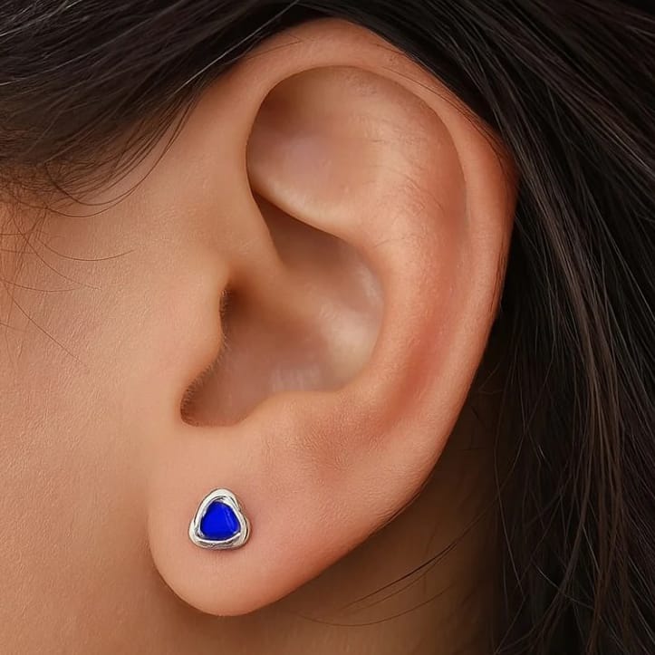 A close-up of an ear wearing a small, triangular earring with a blue stone set in a silver frame.