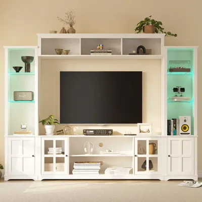 A white entertainment center with shelves and cabinets, holding a flat-screen TV, books, decor items, speakers, and a record player. Soft lighting highlights the shelves.