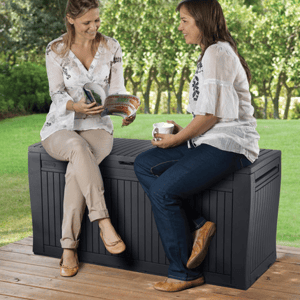 Two women sitting on a black outdoor storage bench while holding books and a mug.