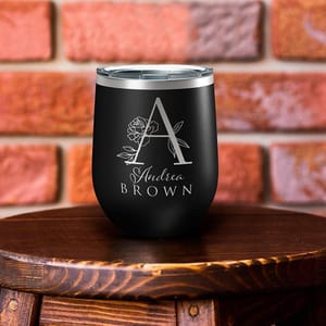 A black stainless steel wine tumbler features an engraved large \“A\“ with a floral design and the name \“Andrea Brown\“ in silver lettering, set against a brick background.