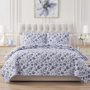 A 3-piece quilt set featuring a blue floral print on a white background, including a quilt and two matching pillow shams. The bed is styled with a tufted headboard, placed in a bright room with white furniture and decor.
