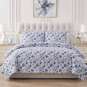 A 3-piece quilt set featuring a blue floral print on a white background, including a quilt and two matching pillow shams. The bed is styled with a tufted headboard, placed in a bright room with white furniture and decor.