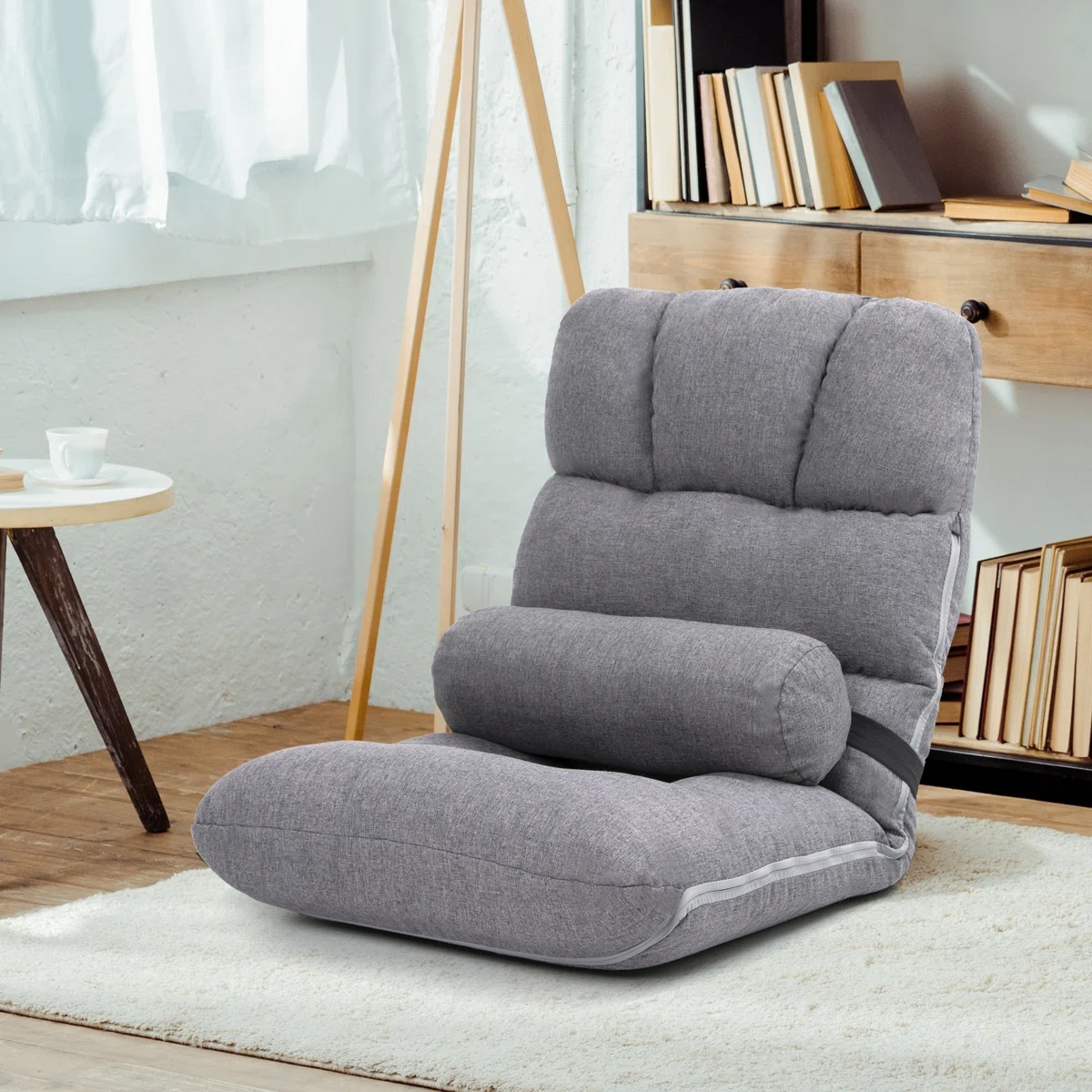A gray, adjustable floor chair with a cushioned backrest and lumbar support pillow, positioned on a light-colored rug beside a coffee table and bookcase.