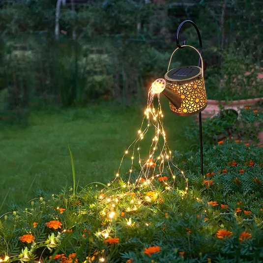 A solar-powered watering can with a decorative cut-out design, hung on a hook, features cascading warm white LED fairy lights that resemble water pouring onto flowers below.