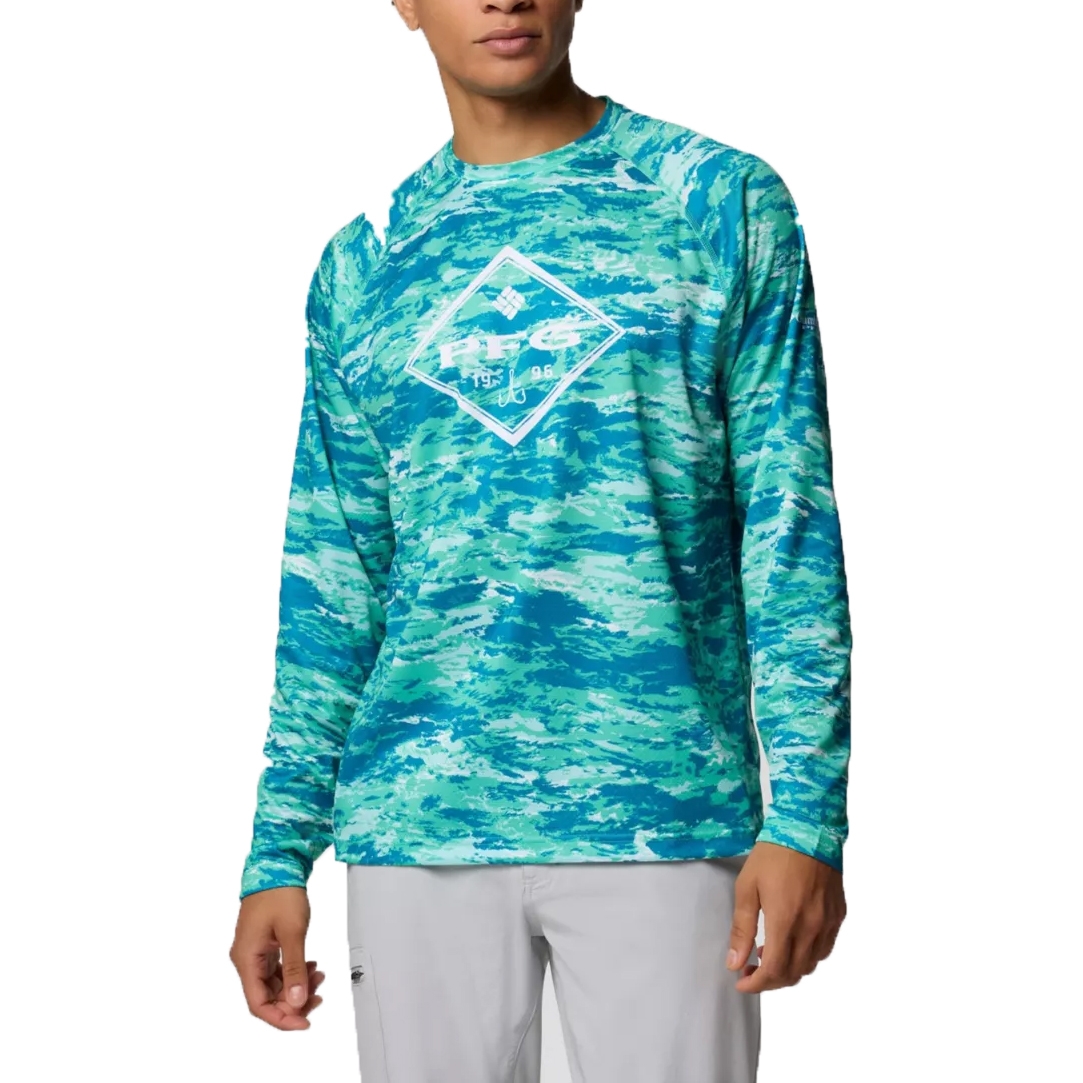 A man wears a long-sleeve shirt featuring a blue and green camouflage pattern with a central diamond-shaped logo. He pairs it with light gray pants.