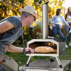 A person using a portable outdoor pizza oven with a pizza inside. Two other people are in the background in a garden setting.