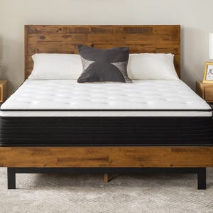 A bed with a wooden headboard features a white and black mattress, flanked by two wooden nightstands with lamps and framed art. A dark decorative pillow is centered on the bed.