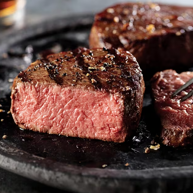 A medium-rare steak sits on a dark plate, seasoned with pepper and lightly seared, with a piece cut to show the pink interior.
