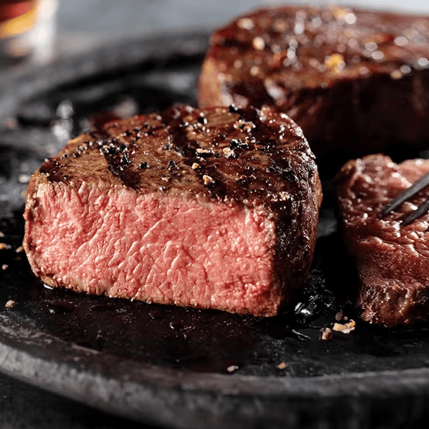 A medium-rare steak sits on a dark plate, seasoned with pepper and lightly seared, with a piece cut to show the pink interior.
