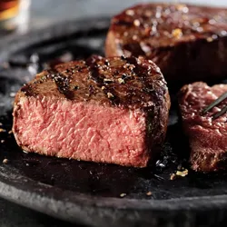 A medium-rare steak sits on a dark plate, seasoned with pepper and lightly seared, with a piece cut to show the pink interior.