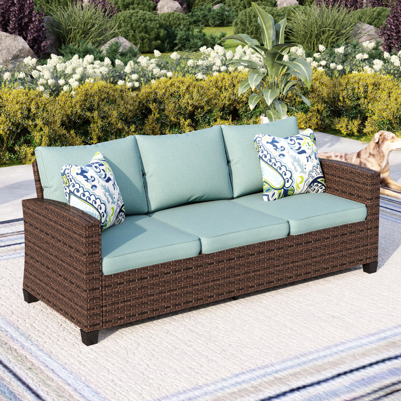 Outdoor wicker sofa with teal cushions and patterned throw pillows, set in a garden. A dog is visible to the side.
