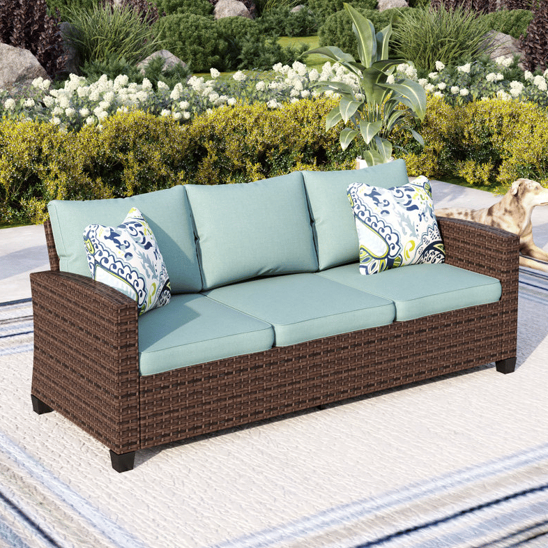 Outdoor wicker sofa with teal cushions and patterned throw pillows, set in a garden. A dog is visible to the side.