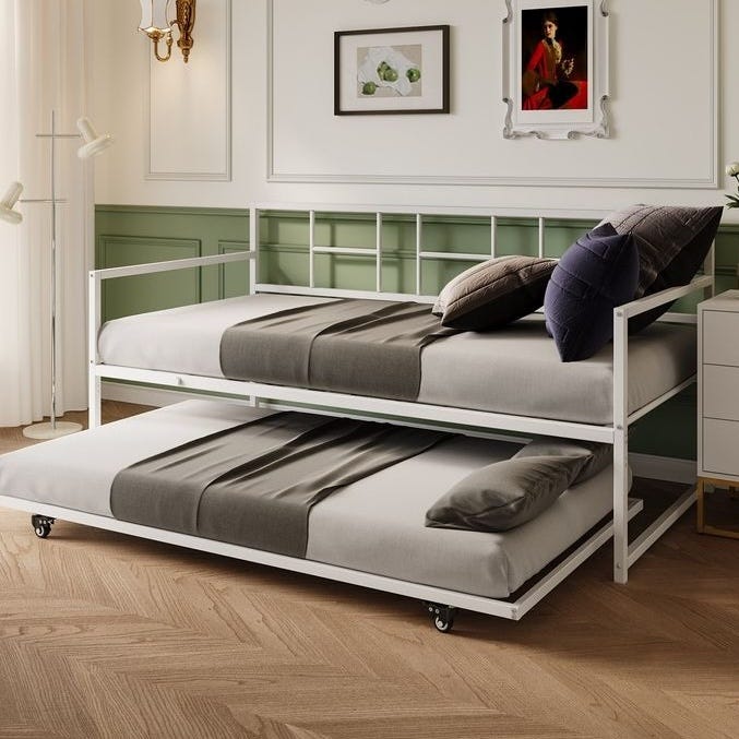 A white metal daybed with a pull-out trundle bed is shown, both featuring grey bedding and pillows. The room is decorated in a classic style with light green paneling, framed artwork, and warm wooden floors.