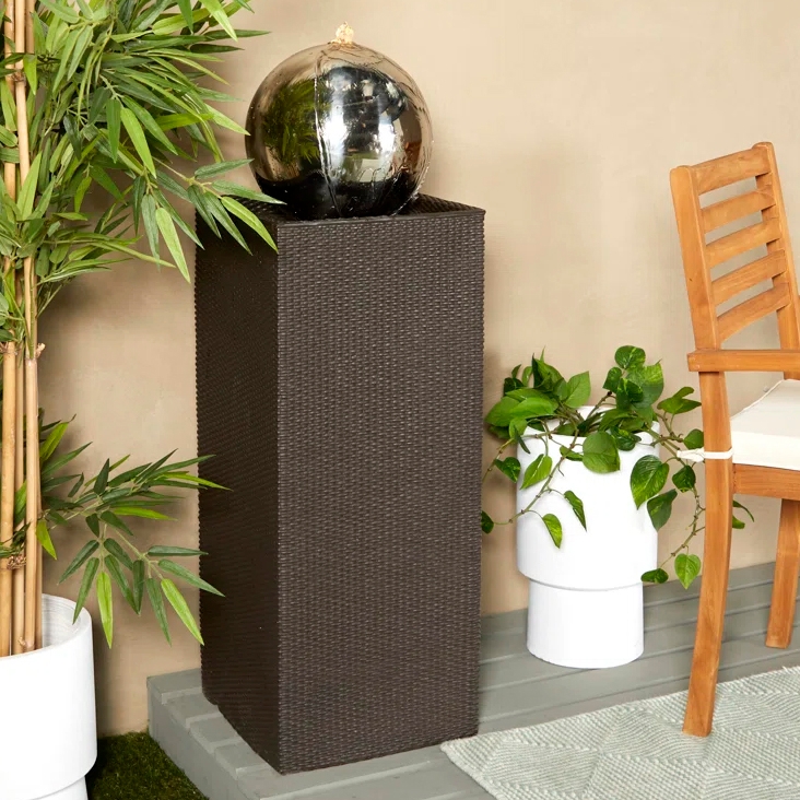 A weather-resistant floor fountain features a brown woven pedestal with a reflective spherical top, suitable for outdoor use. The design is complemented by plants and a wooden chair, providing a serene garden aesthetic.