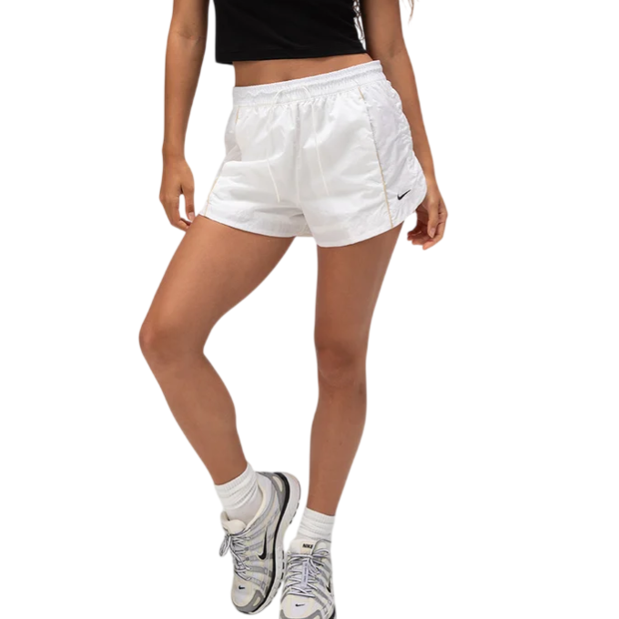 A woman is wearing a black cropped T-shirt with \“NXE\“ lettering, white athletic shorts, Nike socks, and white Nike sneakers.