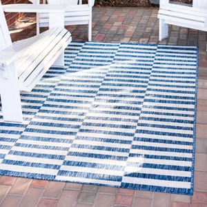 A 7' x 10' area rug features a blue and white striped checkered pattern, placed on a brick patio surrounded by white outdoor chairs.