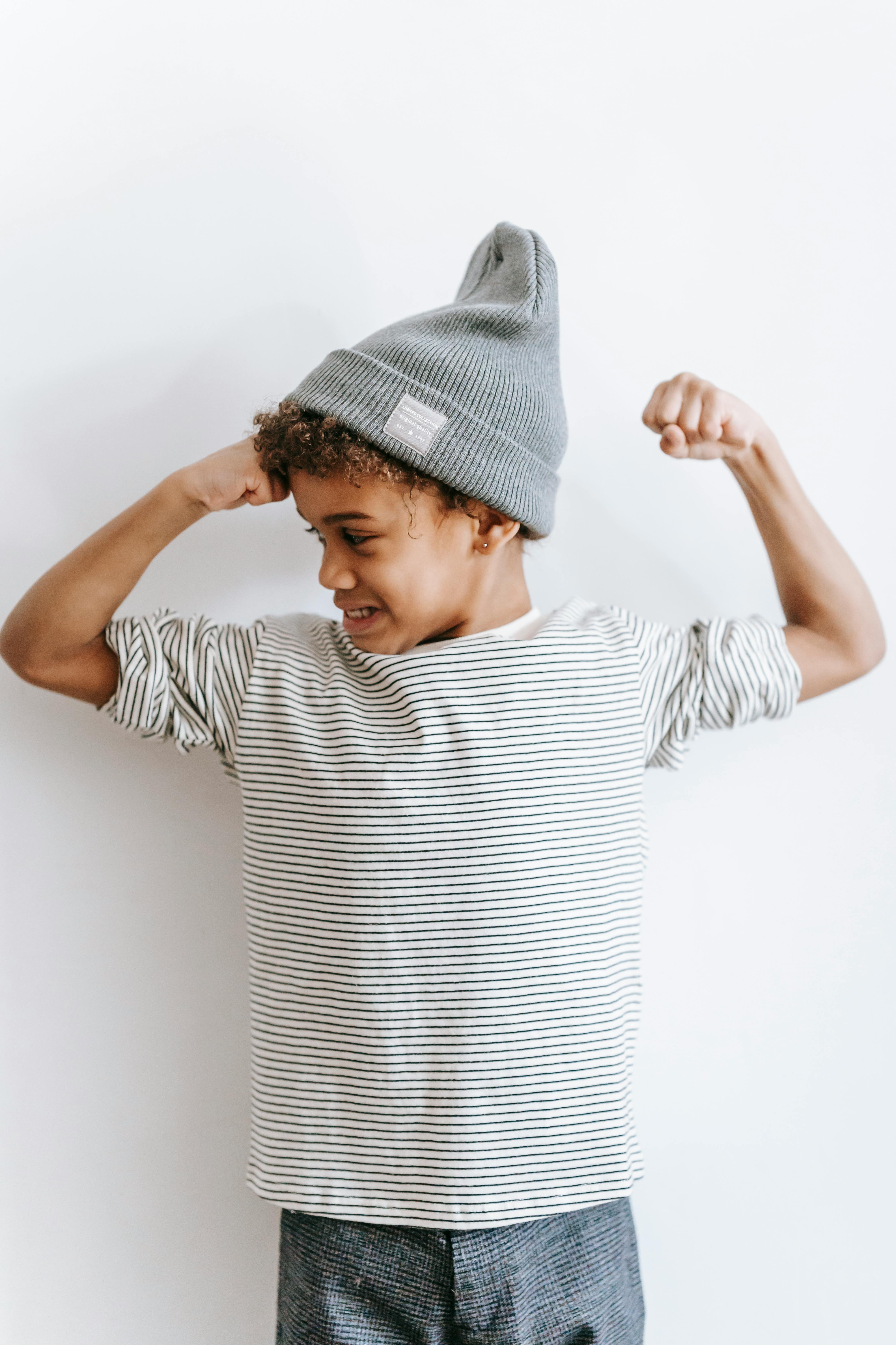 A young person flexing an arm muscle, wearing a gray knit beanie and a white and black striped long-sleeve shirt.