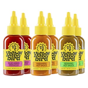 Six bottles of Yellowbird Hot Sauce in various flavors, including Blue Agave Sriracha, Jalapeño, Habanero, and Serrano, with bright yellow labels and yellow squeeze tops.