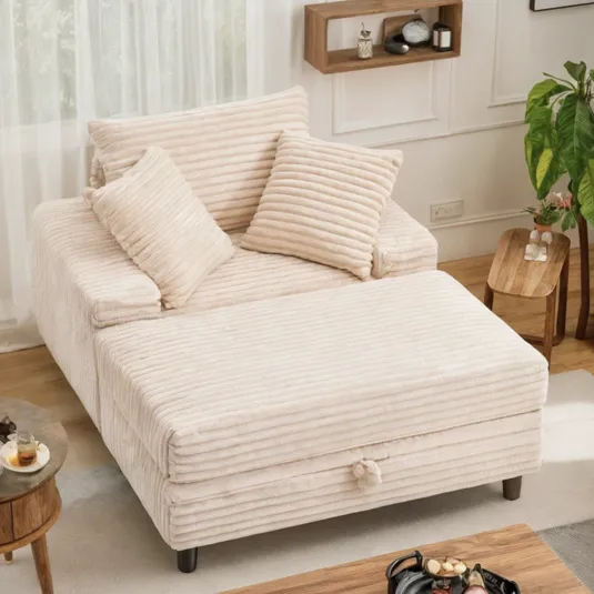 A cream-colored, ribbed fabric sofa with two pillows, featuring an extended chaise. It is placed in a living room setting with wooden and modern decor elements.