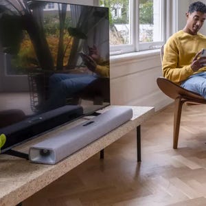 A man sitting on a chair using a smartphone with a soundbar and a flat-screen television on a media console in a living room.