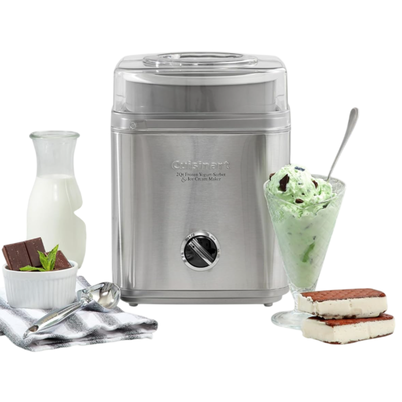 A stainless steel ice cream maker sits on a kitchen counter alongside a glass of mint chocolate chip ice cream, a ramekin with chocolate pieces, and a small carafe of milk.
