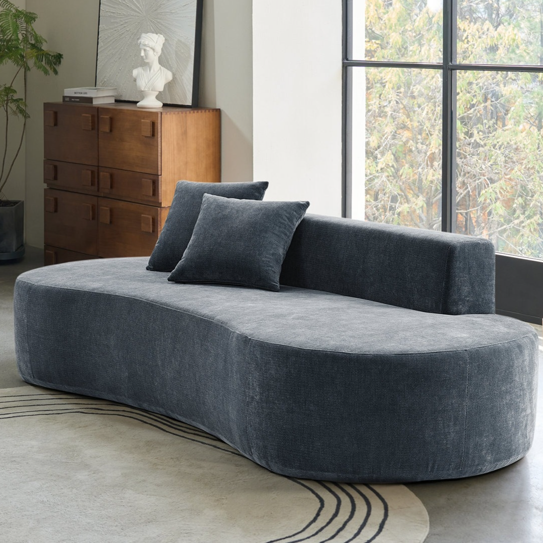 Curved dark gray sofa with two matching pillows set in a room with a large window, wooden dresser with a bust and book on top, geometric patterned rug, and a wooden side table with a lamp.