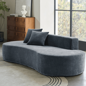 Curved dark gray sofa with two matching pillows set in a room with a large window, wooden dresser with a bust and book on top, geometric patterned rug, and a wooden side table with a lamp.