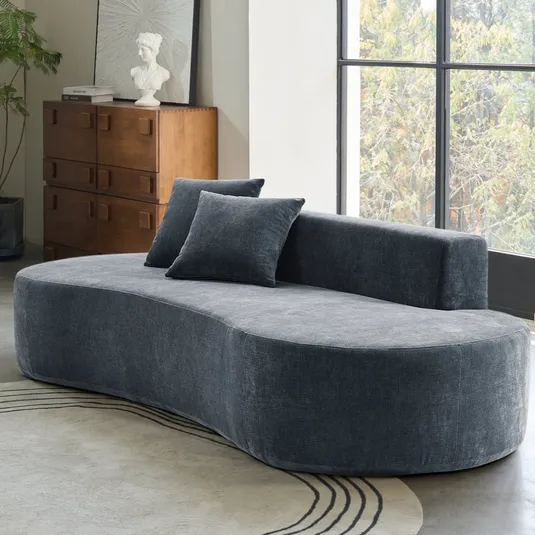 Curved dark gray sofa with two matching pillows set in a room with a large window, wooden dresser with a bust and book on top, geometric patterned rug, and a wooden side table with a lamp.