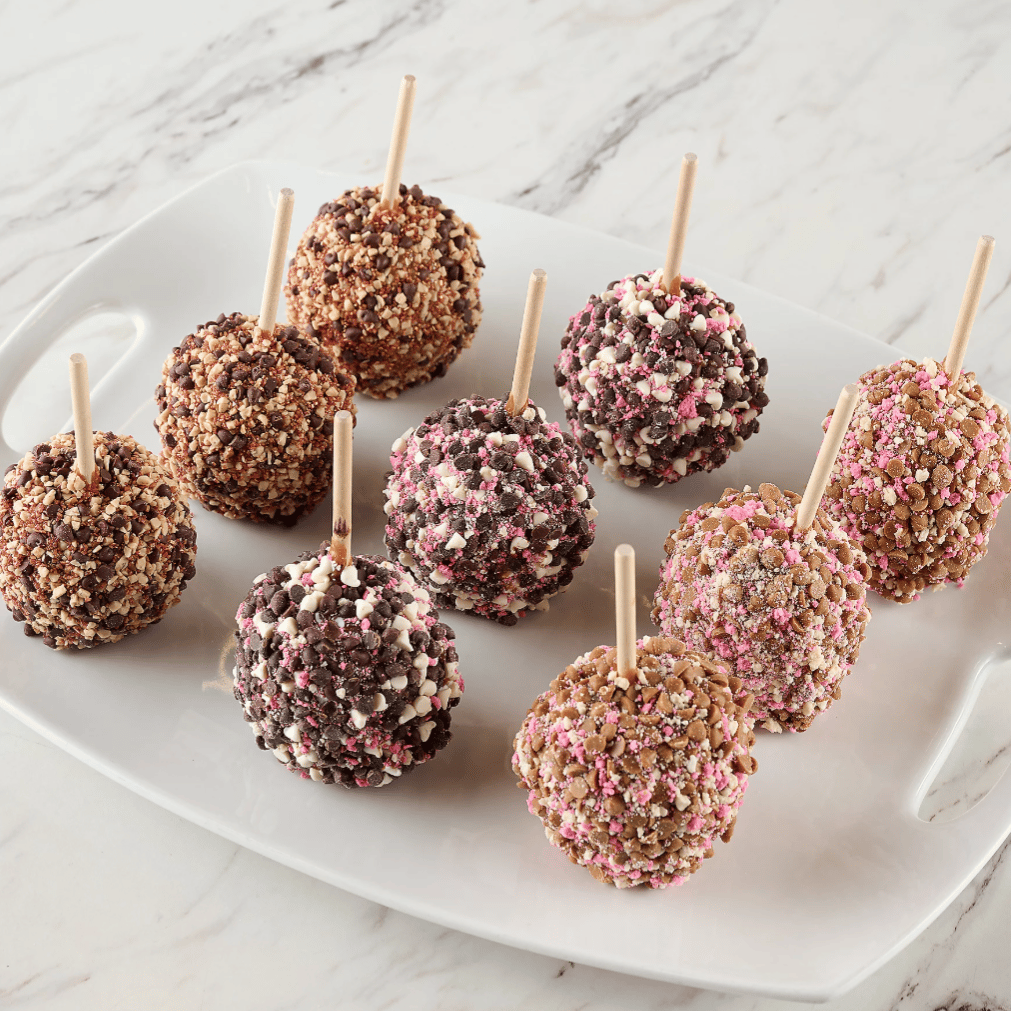 A plate of candy apples coated with different toppings, including nuts and colorful sprinkles, each with a wooden stick for easy handling.
