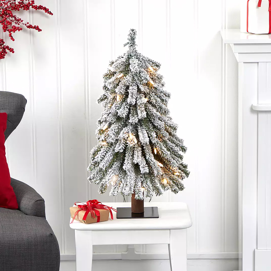A small, snowy artificial Christmas tree with integrated lights, standing 24 inches tall on a table. It features frosted branches for a wintery look and includes a gift box with a red ribbon.