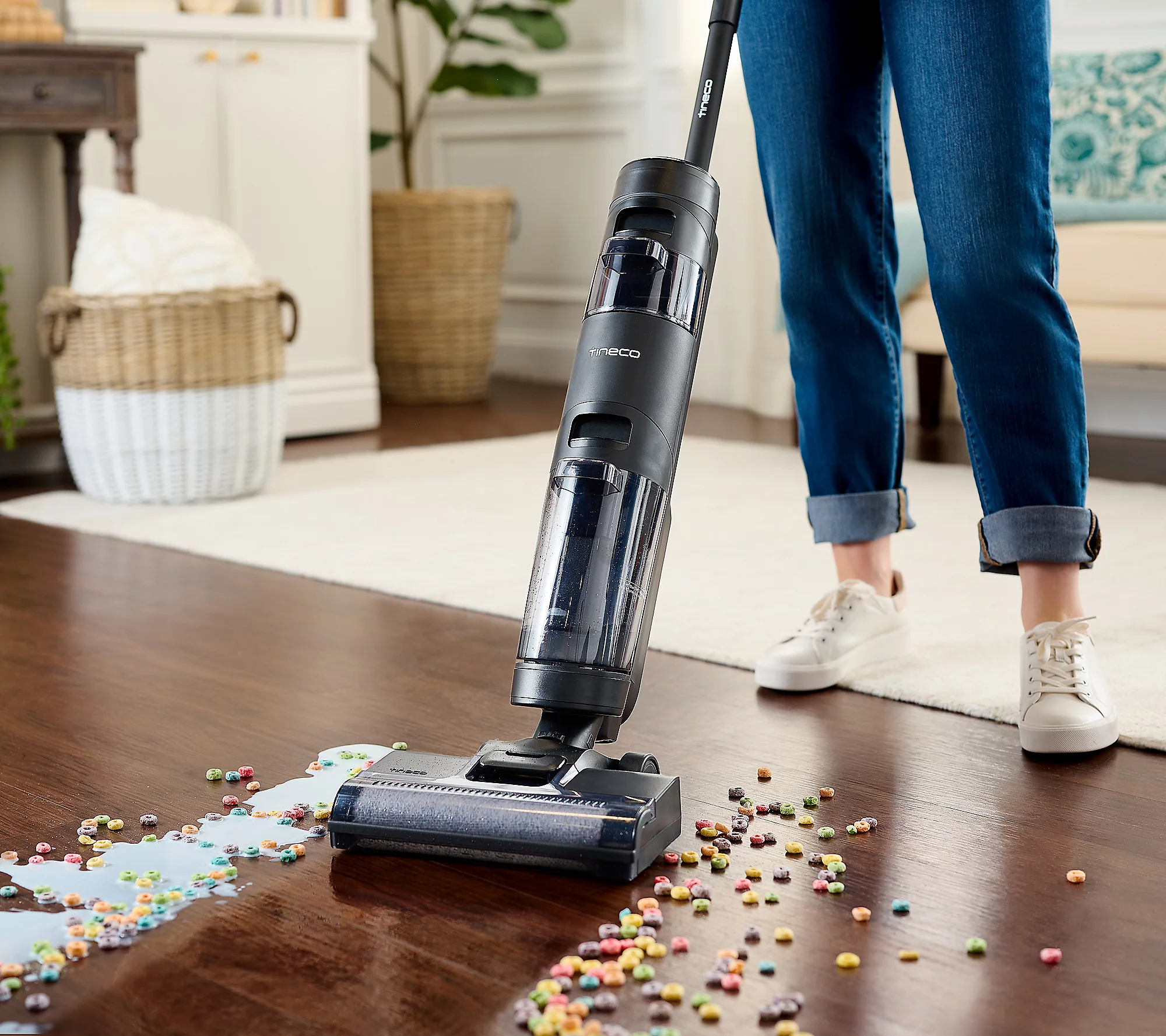A cordless stick vacuum cleaner is being used to pick up colorful cereal scattered on a wooden floor.
