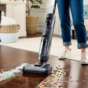 A cordless stick vacuum cleaner is being used to pick up colorful cereal scattered on a wooden floor.