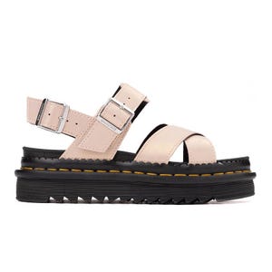 A beige Dr. Martens sandal with two adjustable buckle straps and a thick black rubber sole featuring a yellow stitching detail. The sole has a textured, rugged design for enhanced grip.