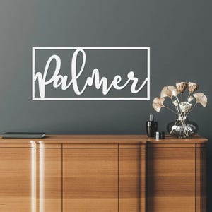 A wooden sideboard with a vase of flowers and cosmetics on top, and a personalized 'Palmer' wall decal above it.