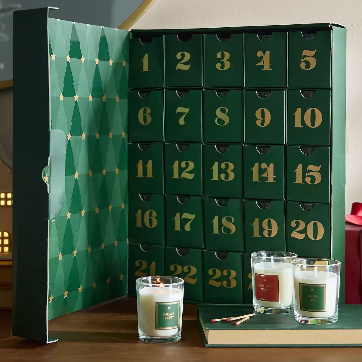 Green advent calendar with 25 numbered drawers and three scented candles labeled \“Balsam Forest,\“ \“Peppermint Candy,\“ and \“Snow Day,\“ alongside matches on a wooden surface.