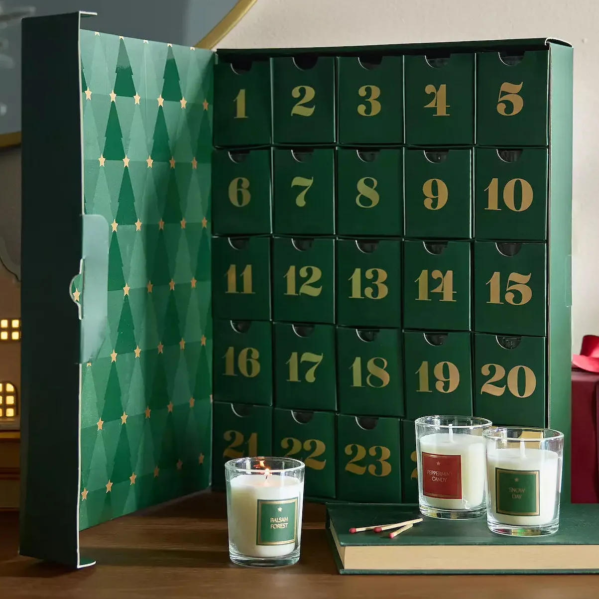 Green advent calendar with 25 numbered drawers and three scented candles labeled \“Balsam Forest,\“ \“Peppermint Candy,\“ and \“Snow Day,\“ alongside matches on a wooden surface.