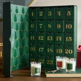 Green advent calendar with 25 numbered drawers and three scented candles labeled \“Balsam Forest,\“ \“Peppermint Candy,\“ and \“Snow Day,\“ alongside matches on a wooden surface.