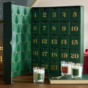 Green advent calendar with 25 numbered drawers and three scented candles labeled \“Balsam Forest,\“ \“Peppermint Candy,\“ and \“Snow Day,\“ alongside matches on a wooden surface.