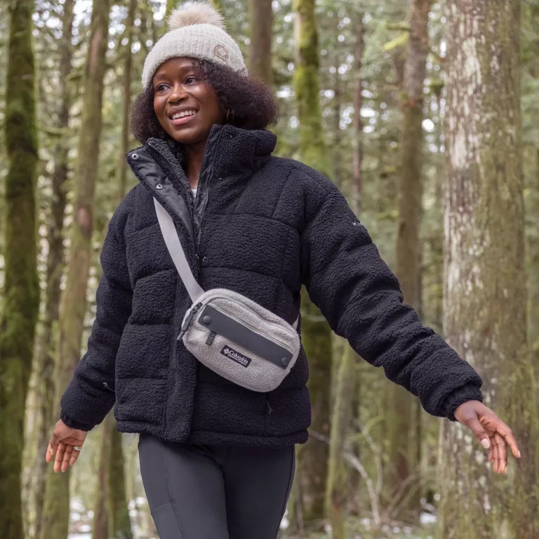 A woman wearing a black puffer jacket, beige beanie, and sporting a grey crossbody bag stands in a forest.